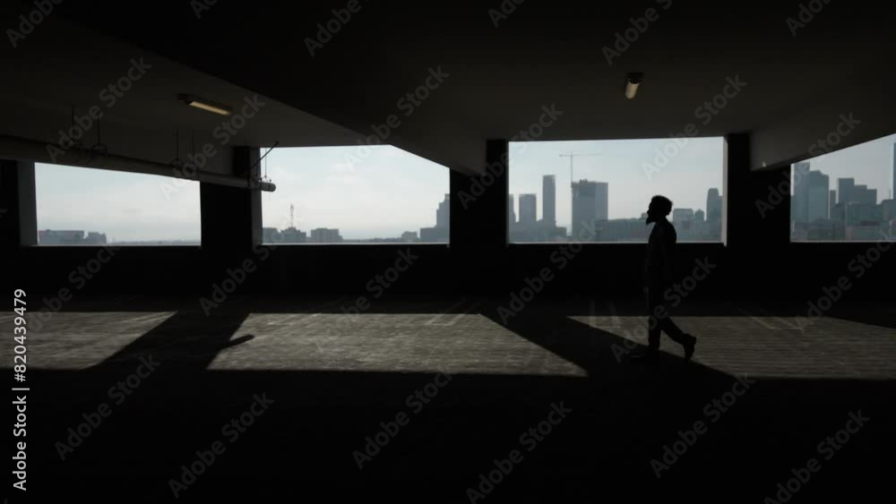 Silhouette of a businessman walks in a dark urban parking garage, with the city skyline in the background. The shadowy setting creates an air of mystery and a corporate ambiance. Slow Motion. Silhouette of a businessman walks in a dark urban parking garage, with the city skyline in the background. The shadowy setting creates an air of mystery and a corporate ambiance. Slow Motion.