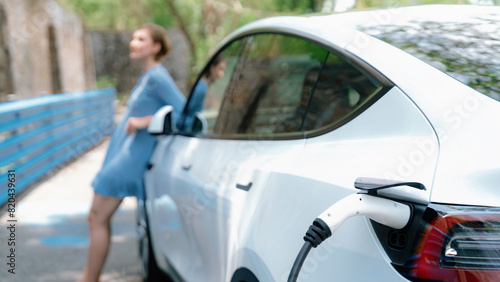 Focus EV charger by electric charging station recharging car on blurred background of beautiful young woman checking car's battery from smartphone. Eco-friendly travel with EV car. Panorama Perpetual