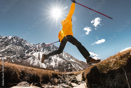 Young woman hiker jumps with hiking poles over the gap with stones against sun and mountain