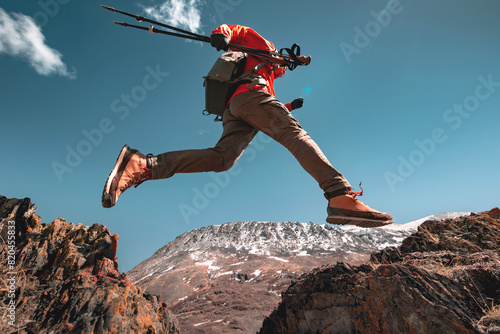Man hiker with hiking poles and backpack jumps over the hole against clear blue sky and mountains