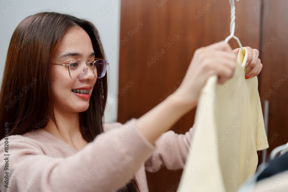 Young Asian girl with long brown hair, wearing pink sweater and glasses, smiling while holding yellow sweater on hanger. happy and fresh expression, emphasizing personal style and fashion choices. Young Asian girl with long brown hair, wearing pink sweater and glasses, smiling while holding yellow sweater on hanger. happy and fresh expression, emphasizing personal style and fashion choices.