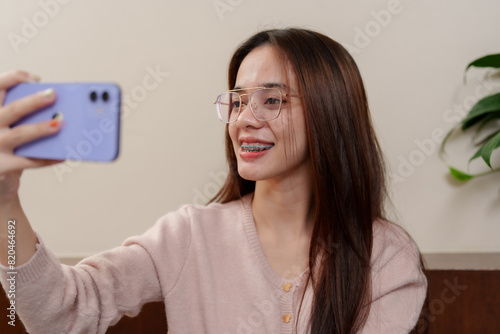 Asian teenage girl with long brown hair, wearing pink sweater and glasses, smiling while taking selfie with smartphone. happy and fresh expression, emphasizing self-expression and connection.