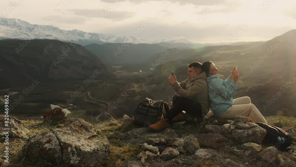 A couple of tourists, a man and a woman, relax on the top of a mountain at sunset and look at their phones. Tourists correspond on social networks. A couple of tourists, a man and a woman, relax on the top of a mountain at sunset and look at their phones. Tourists correspond on social networks.