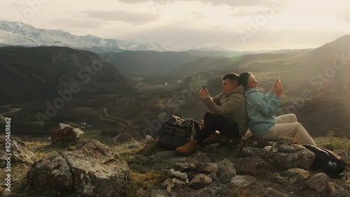 A couple of tourists, a man and a woman, relax on the top of a mountain at sunset and look at their phones. Tourists correspond on social networks. photo