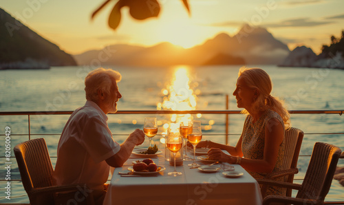 Elderly couple enjoying a romantic sunset dinner on a scenic waterfront patio with mountains in the background.