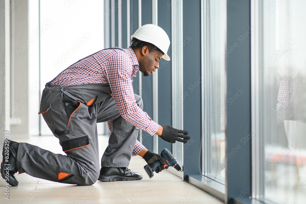 Low Angle View Of A Young African Repairman In Overalls Installing Window Low Angle View Of A Young African Repairman In Overalls Installing Window