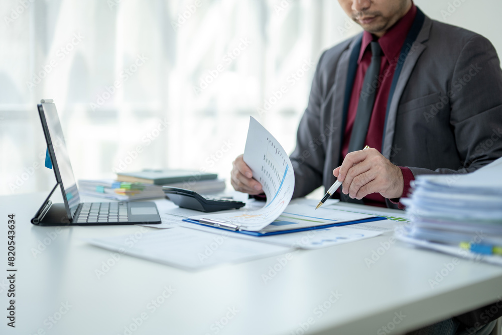 A man in a suit is sitting at a desk with a laptop and a stack of papers A man in a suit is sitting at a desk with a laptop and a stack of papers