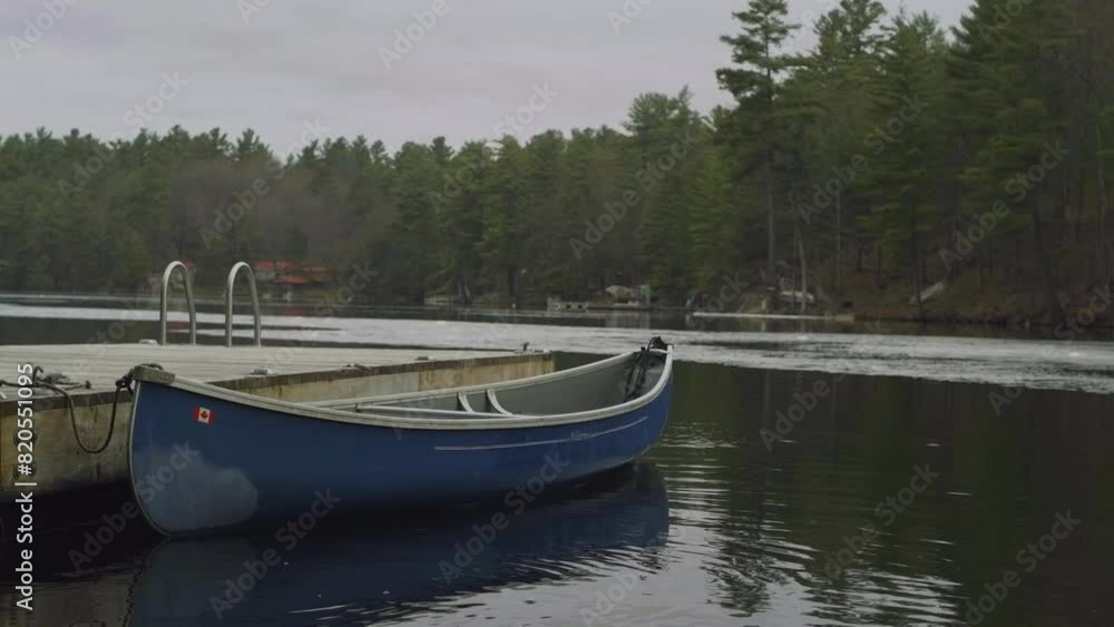 A blue canoe is docked at a wooden pier on a calm lake, surrounded by tall pine trees and a serene natural landscape, under an overcast sky. A blue canoe is docked at a wooden pier on a calm lake, surrounded by tall pine trees and a serene natural landscape, under an overcast sky.