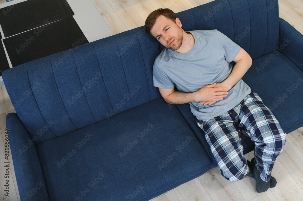 Fototapeta premium Guy with a beard feeling unwell with a stomach ache while sitting in a couch at home