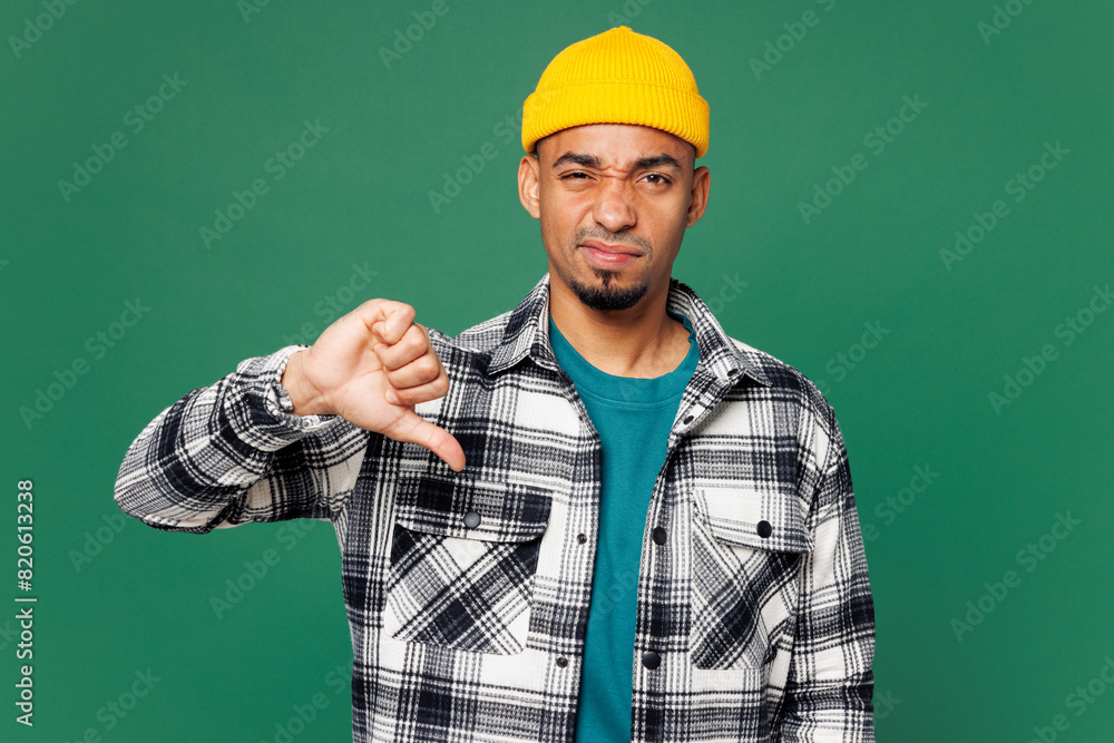 Young sad dissatisfied man of African American ethnicity he wear shirt blue t-shirt yellow hat showing thumb down dislike gesture isolated on plain green background studio portrait. Lifestyle concept. Young sad dissatisfied man of African American ethnicity he wear shirt blue t-shirt yellow hat showing thumb down dislike gesture isolated on plain green background studio portrait. Lifestyle concept.