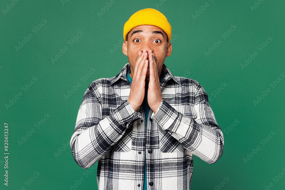 Young shocked surprised man of African American ethnicity wear shirt blue t-shirt yellow hat look camera cover mouth with hands isolated on plain green background studio portrait. Lifestyle concept Young shocked surprised man of African American ethnicity wear shirt blue t-shirt yellow hat look camera cover mouth with hands isolated on plain green background studio portrait. Lifestyle concept