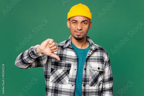 Young sad dissatisfied man of African American ethnicity he wear shirt blue t-shirt yellow hat showing thumb down dislike gesture isolated on plain green background studio portrait. Lifestyle concept.