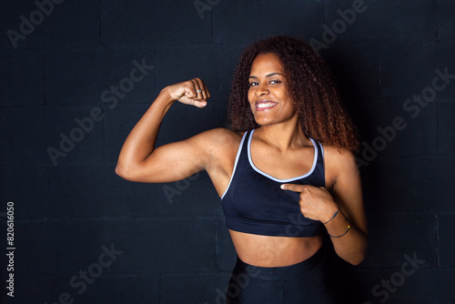 Mujer deportista señalando su biceps. Biceps de mujer. Chica en gimnasio. Fondo negro. Mujer con ropa de deporte. Chica sonriente. Estilo de vida saludable