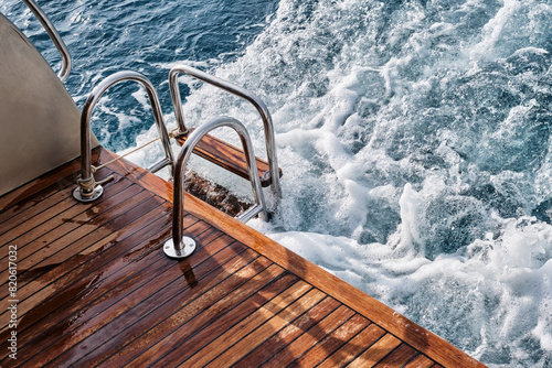 Fotografie Back teak wooden deck and metal ladder of a of motor yacht, waves and sea splashes and foam