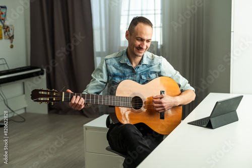 Portrait of man taking online guitar lesson looking at laptop screen. Retired male learning to play guitar watching webinar on computer at home