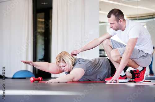 Overweight woman exercising on gym mat in gym. Personal trainer couching her and helping her.