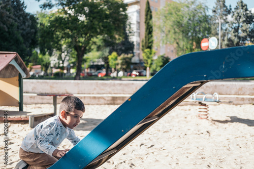 Young autistic child playing on a slide at the park. Little boy enjoys a sunny day climbing up a blue slide in an outdoor playground
