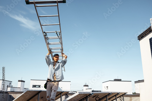 Man carries ladder during solar panels installation on a rooftop of a private house on a sunny day. Wide angle view from below. Concept of maintenance and installing solar power station