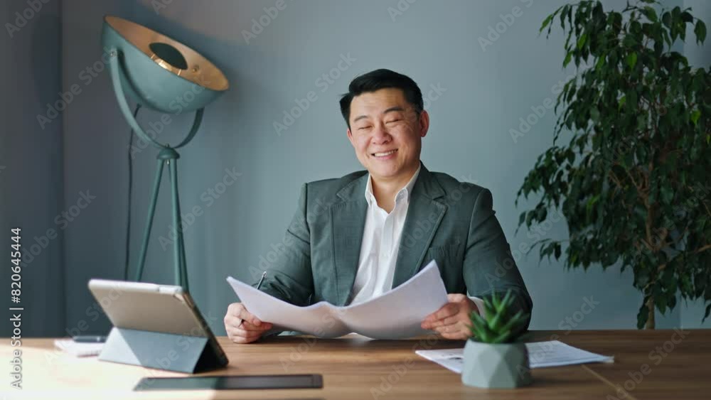 Smiling japanese male clutching documents in hands and gazing confidently into camera. Successful young manager sitting in front of modern tablet in office environment. Concept of occupation. Smiling japanese male clutching documents in hands and gazing confidently into camera. Successful young manager sitting in front of modern tablet in office environment. Concept of occupation.