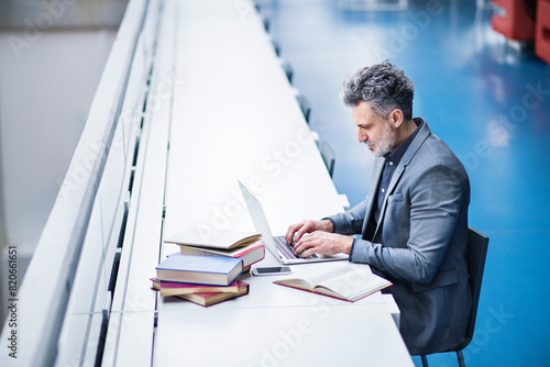 Handsome businessman working on laptop, searching for informations in book. Traditional information source in modern office space. photo