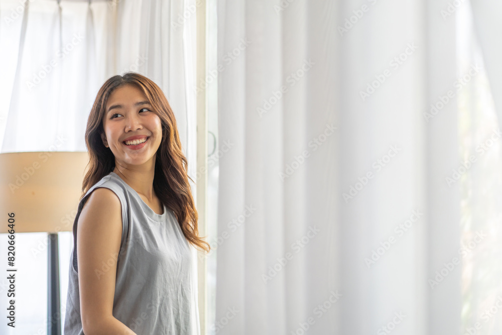 Happy young beautiful pretty asian woman waking up and opening window curtains.Girl feeling comfortable and relaxed breathing fresh air in morning in bedroom and copy space banner at home