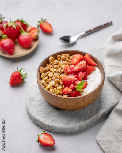Natural yogurt with granola and strawberries in a wooden bowl on a marble board on a light background with fresh berries.