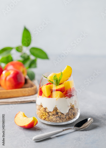 Yogurt parfait with peach and granola in a glass on a light background with fresh fruits. Healthy eating concept.