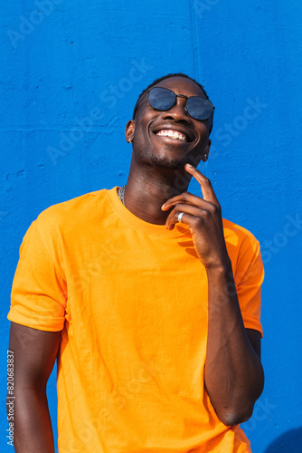 Young cheerful African American male in bright yellow t shirt and sunglasses looking upwards standing against blue wall photo