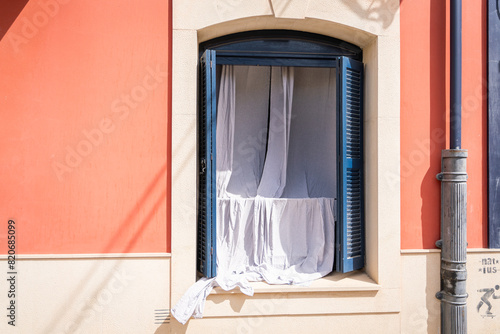 White curtains hang from a window in an old  building.  Window with blue shutters on a building withwhite and  pink facade