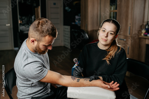 Young woman professional tattoo artist in black gloves making a rose tattoo on a man's forearm.