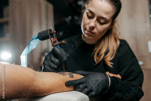 Young woman professional tattoo artist in black gloves making a rose tattoo on a man's forearm.