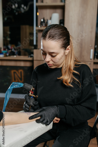 Close up young woman professional tattoo artist in black gloves making a  tattoo on a woman's  forearm