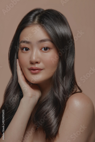 Beautiful brunette asian woman with a clean healthy skin touching her face over beige background, vertical close up shot.