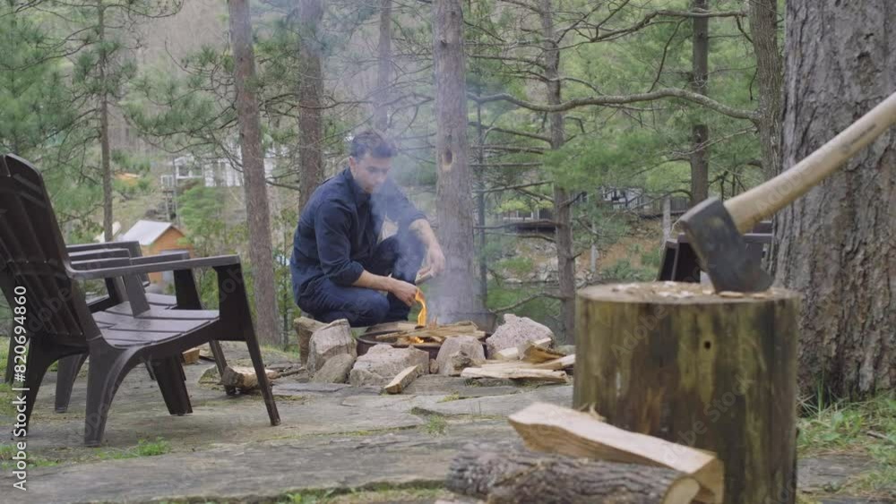 A man is starting a fire in an outdoor fire pit, surrounded by trees. An axe is embedded in a log nearby, with chairs set up around the fire pit, creating a cozy and rustic outdoor scene. A man is starting a fire in an outdoor fire pit, surrounded by trees. An axe is embedded in a log nearby, with chairs set up around the fire pit, creating a cozy and rustic outdoor scene.