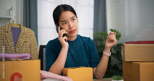 Portrait of Young asian woman business owner sitting at desk talking with customer on smartphone while packing for shipping, Online Shopping