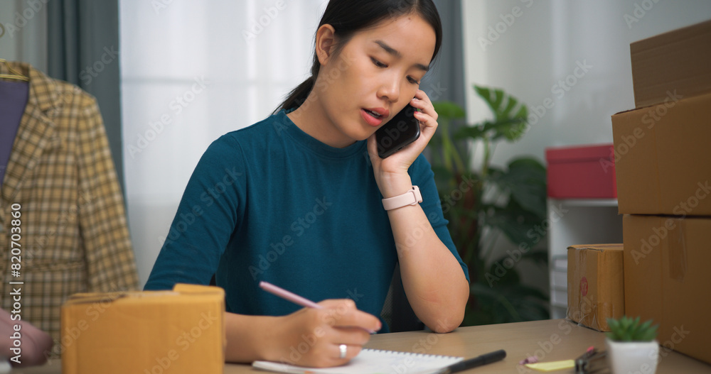 Portrait of Young asian woman business owner sitting at desk use laptop while check orders and write into book, Online Shopping Portrait of Young asian woman business owner sitting at desk use laptop while check orders and write into book, Online Shopping