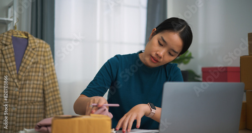 Portrait of  Young asian woman business owner sitting at desk use laptop while check orders and write into book, Online Shopping