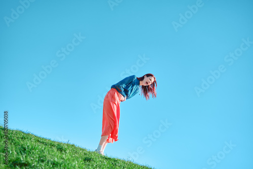 Teenage girl bent over and screaming on a grassy slope against a clear blue sky. Tilted camera angle