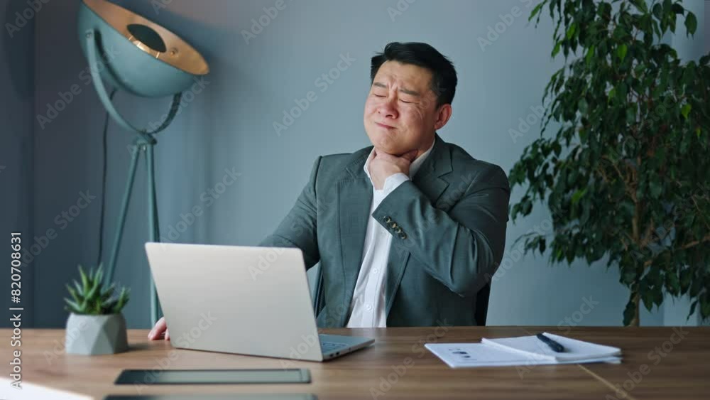 Unhappy japanese gentleman grimacing with throat discomfort during break at a desk adorned with wireless laptop and stylish blue lamp on backdrop. Composed individual gently rubbing throat in office. Unhappy japanese gentleman grimacing with throat discomfort during break at a desk adorned with wireless laptop and stylish blue lamp on backdrop. Composed individual gently rubbing throat in office.