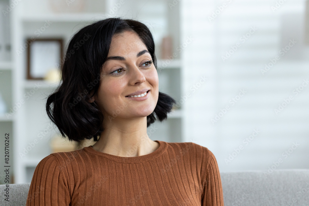 Smiling woman sitting on sofa in living room, looking thoughtful and content Smiling woman sitting on sofa in living room, looking thoughtful and content