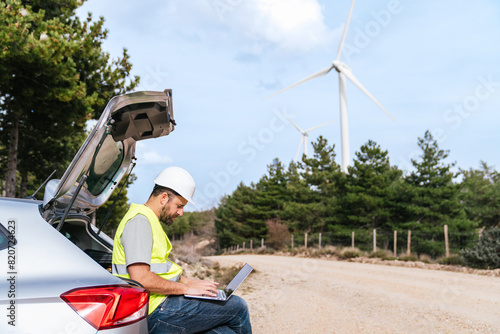 A wind turbine technician sits in the car trunk, wearing a hard hat and reflective vest, working on a laptop.