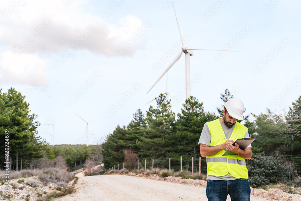 A technician in a high-visibility vest and hard hat takes notes on a clipboard while inspecting wind turbines along a rural pathway. A technician in a high-visibility vest and hard hat takes notes on a clipboard while inspecting wind turbines along a rural pathway.