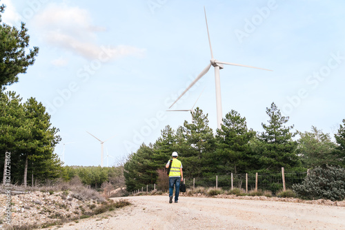 A technician in a reflective vest and hard hat walks on a dirt road, carrying equipment and tools, with wind turbines in the distance.
