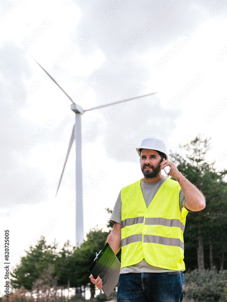 A wind turbine technician talks on the phone while holding a clipboard, wearing a safety vest and helmet, with a wind turbine in the background. A wind turbine technician talks on the phone while holding a clipboard, wearing a safety vest and helmet, with a wind turbine in the background.