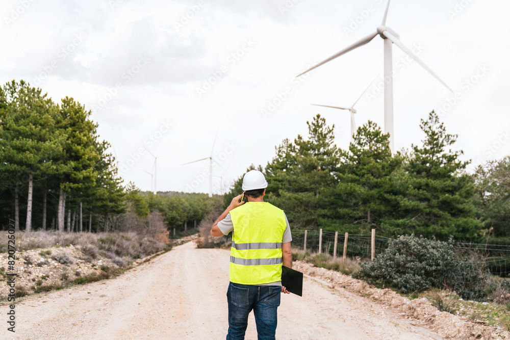 A wind turbine technician stands on a dirt road, talking on the phone and holding a clipboard, with wind turbines and trees in the background. A wind turbine technician stands on a dirt road, talking on the phone and holding a clipboard, with wind turbines and trees in the background.
