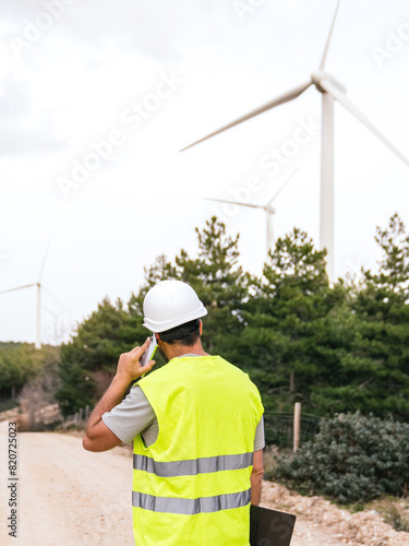 A wind turbine technician speaks on the phone, holding a clipboard, wearing a safety vest and helmet, viewed from the back with turbines in the background.