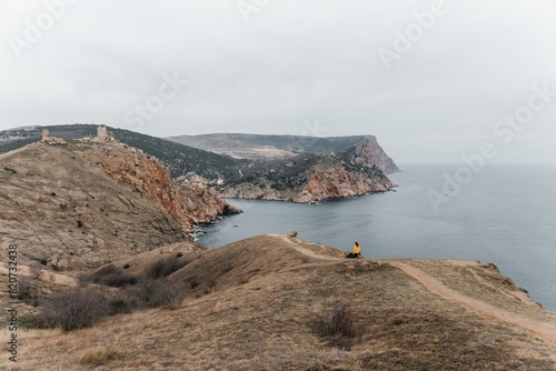 Woman stands on cliff overlooking ocean, cloudy day provides soft lighting. She's wearing a yellow sweater wearing dark pants. Landscape features rugged terrain, calm waters. photo