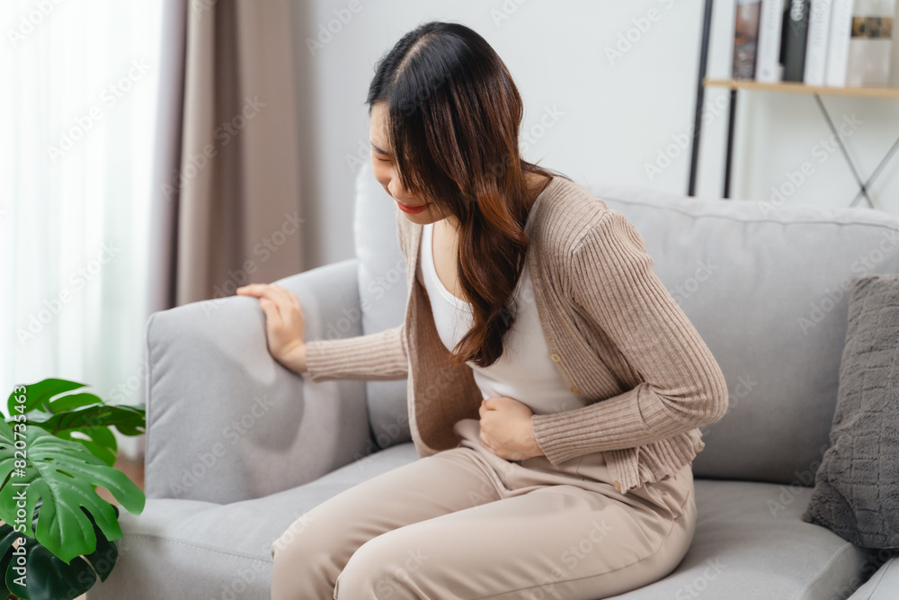 The woman with a stomachache is holding her stomach in pain, sitting on the sofa in the living room. The woman with a stomachache is holding her stomach in pain, sitting on the sofa in the living room.