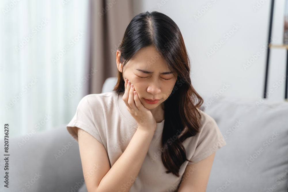 The woman with a toothache is holding her cheek in pain while sitting on the sofa at home. The woman with a toothache is holding her cheek in pain while sitting on the sofa at home.