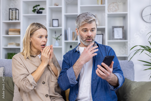Middle-aged couple with shocked expressions, looking at a smartphone while sitting on the couch in a modern living room.